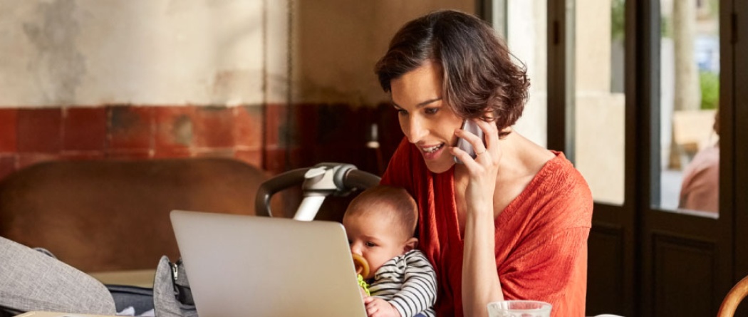 a mother holding her baby in a coffee shop and looking at her laptop